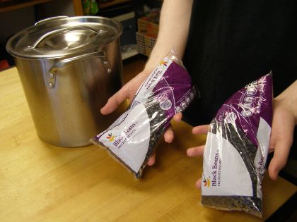 Me holding two one-pound bags of beans over a counter; a metal pot is in the background.